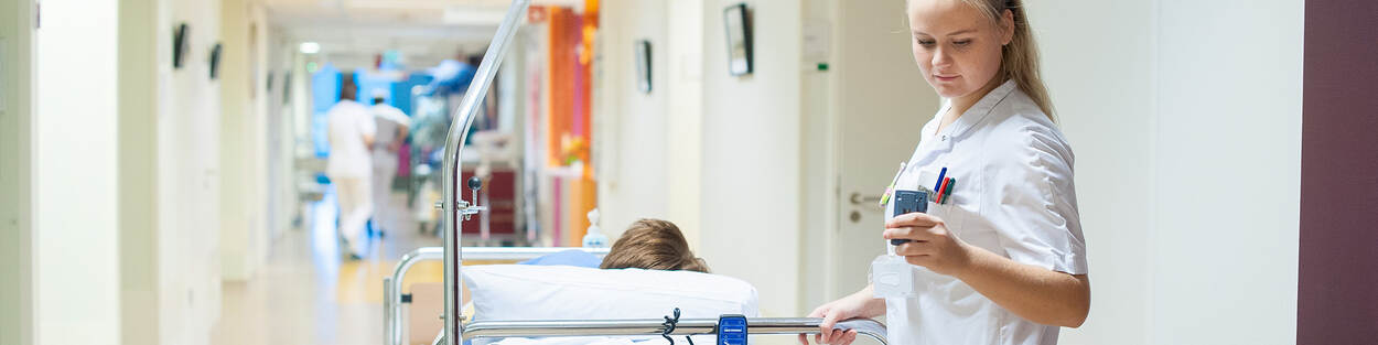 Een verpleegkundige in een ziekenhuis duwt een bed met patiënt en kijkt op haar pieper